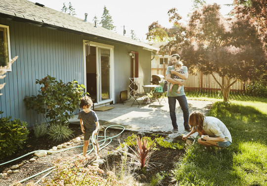 un homme portant un enfant devant une maison et des enfants en train de jouer devant la maison