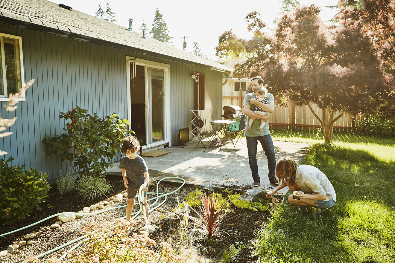 un adulte portant un enfant et 2 enfants dans un jardin devant une maisons. les enfants arronsent les plantes